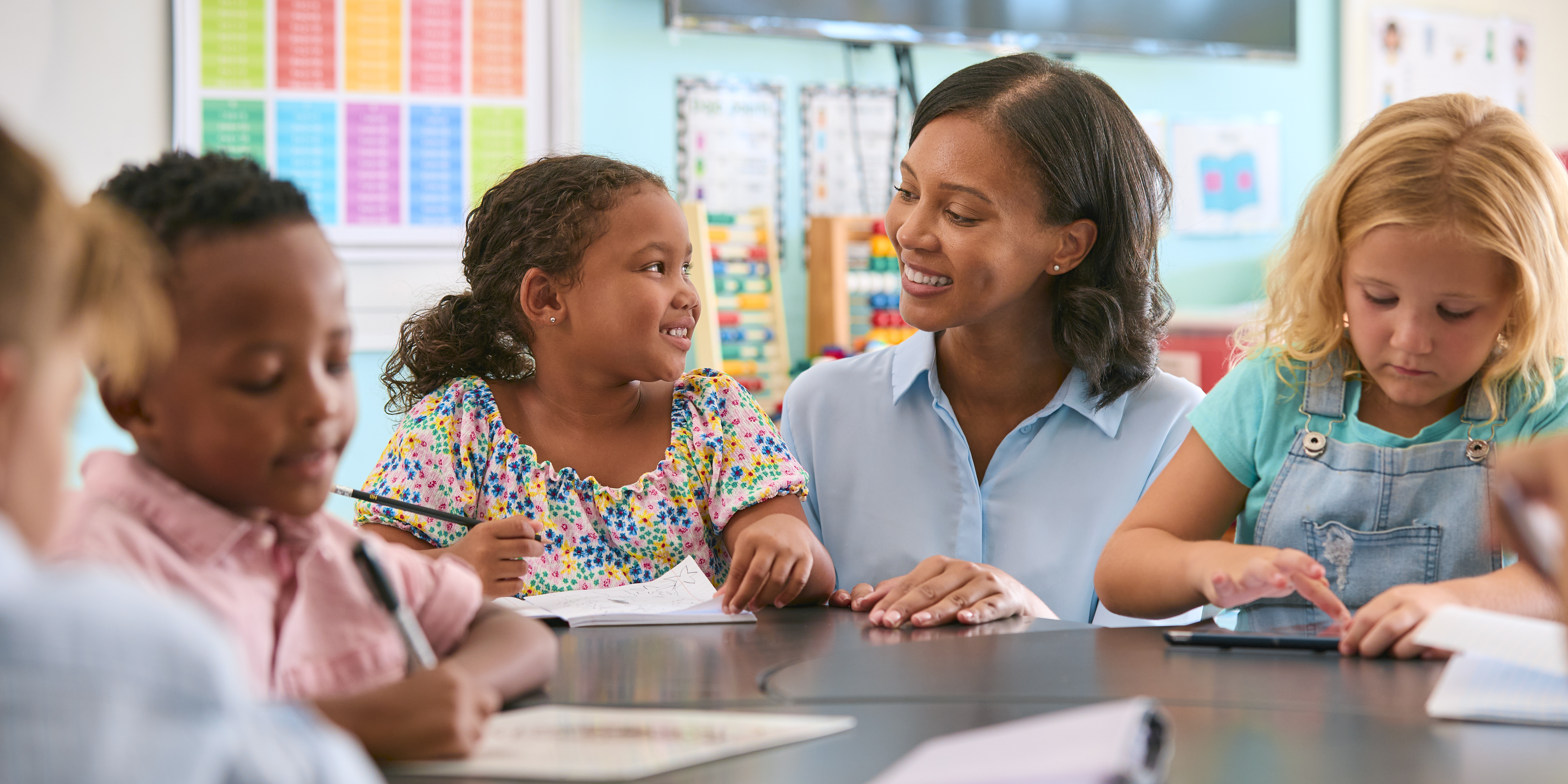 Female Primary Or Elementary School Teacher Helping Students At Desk In Multi-Cultural Class